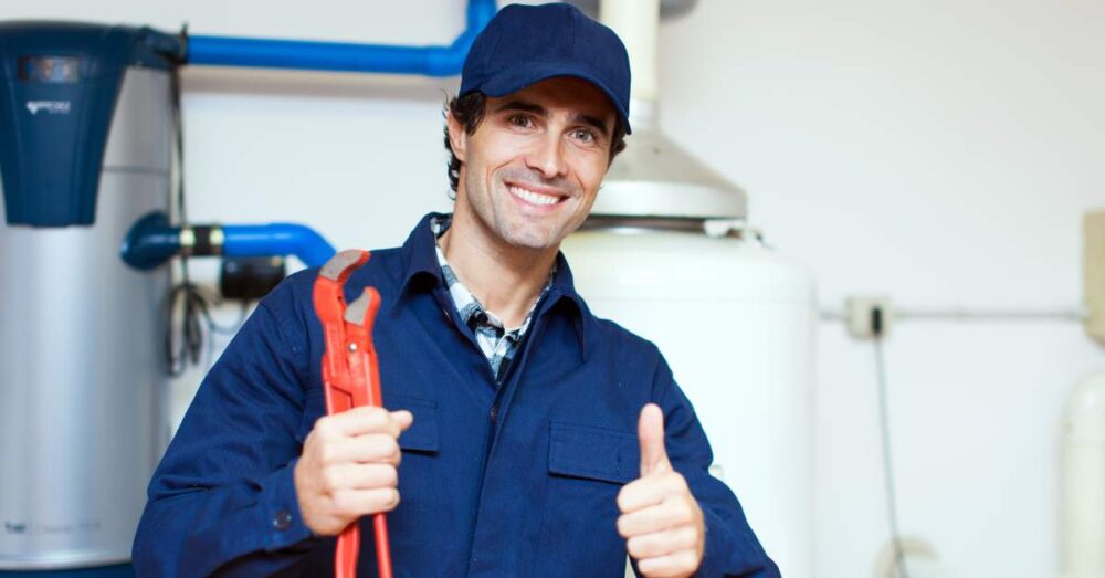 Anlagenmechaniker in Arbeitskleidung mit Rohrzange vor Heizungsanlage, zeigt Daumen hoch