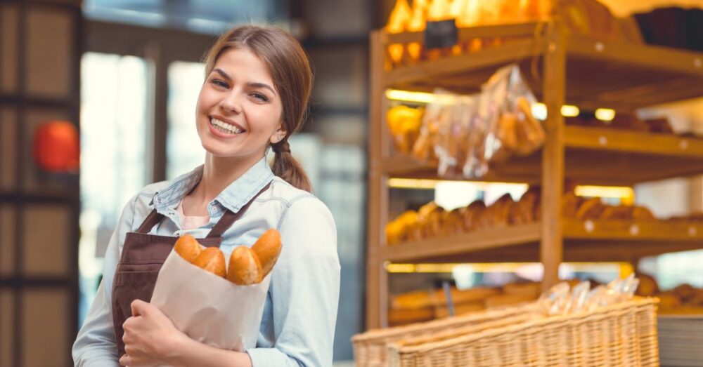 Verkäuferin steht in einer Bäckerei mit einer Brötchentüte in der Hand und lächelt in die Kamera
